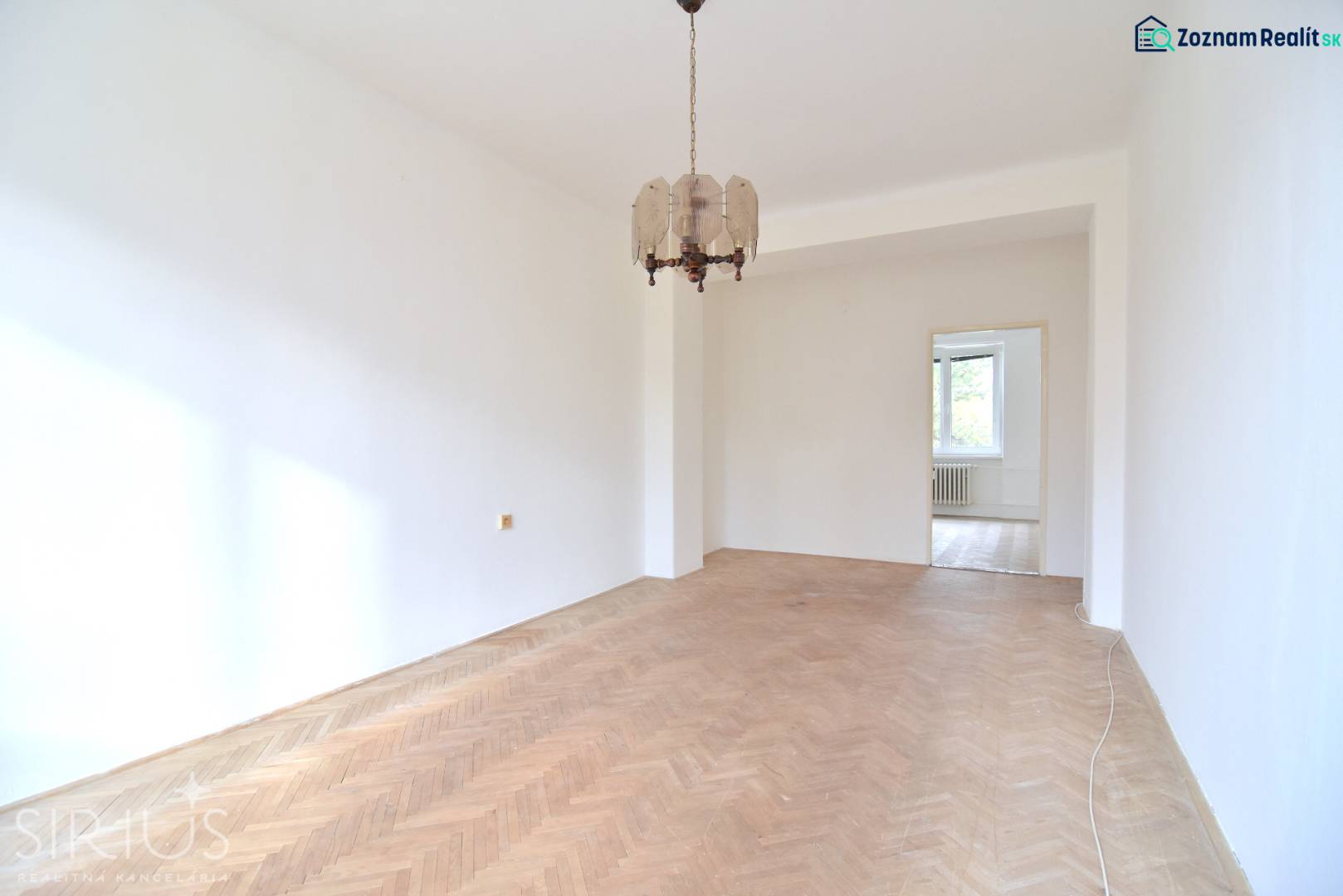 SEČOVSKÁ, 2-i tehlový byt, 56 m2, orientácia V a Z - OBKLOPENÝ ZELEŇOU - Priestranná izba v 2-izbovom byte s podlahou s dreveným dekorom a závesným svietidlom.