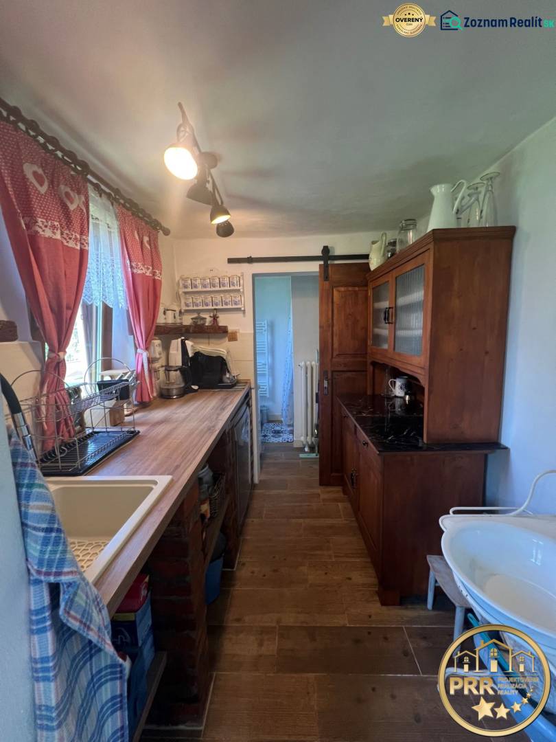 !! NOVÁ CENA !! predaj romantickej chalúpky v Šípkove  - Kuchyňa v drevenej chate s rustikálnym nábytkom, umývadlom a dekoratívnymi záclonami.