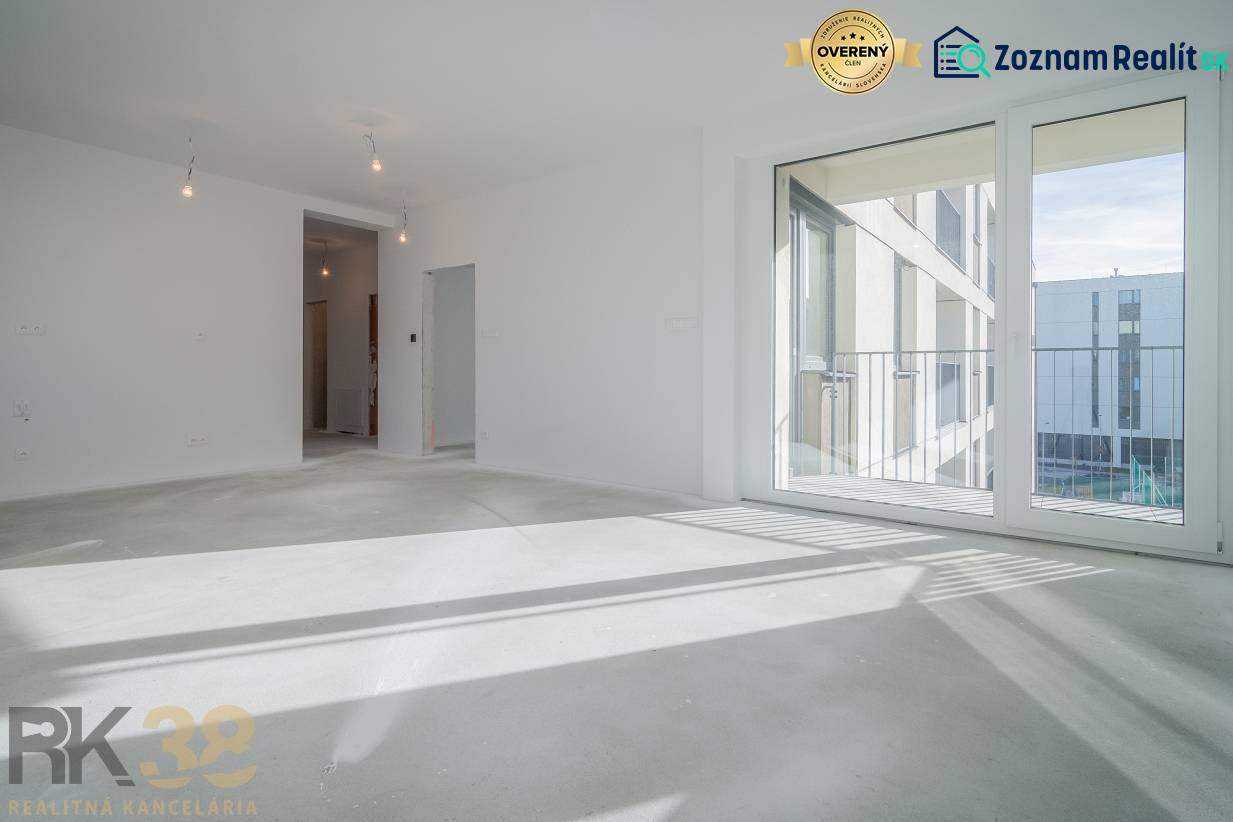 EXKLUZÍVNA PONUKA: Novostavba 3-izb. bytu v projekte Nové Nábrežie - Izba v 3-izbovom byte s veľkým oknom, podlaha s dreveným dekorom, biele steny.