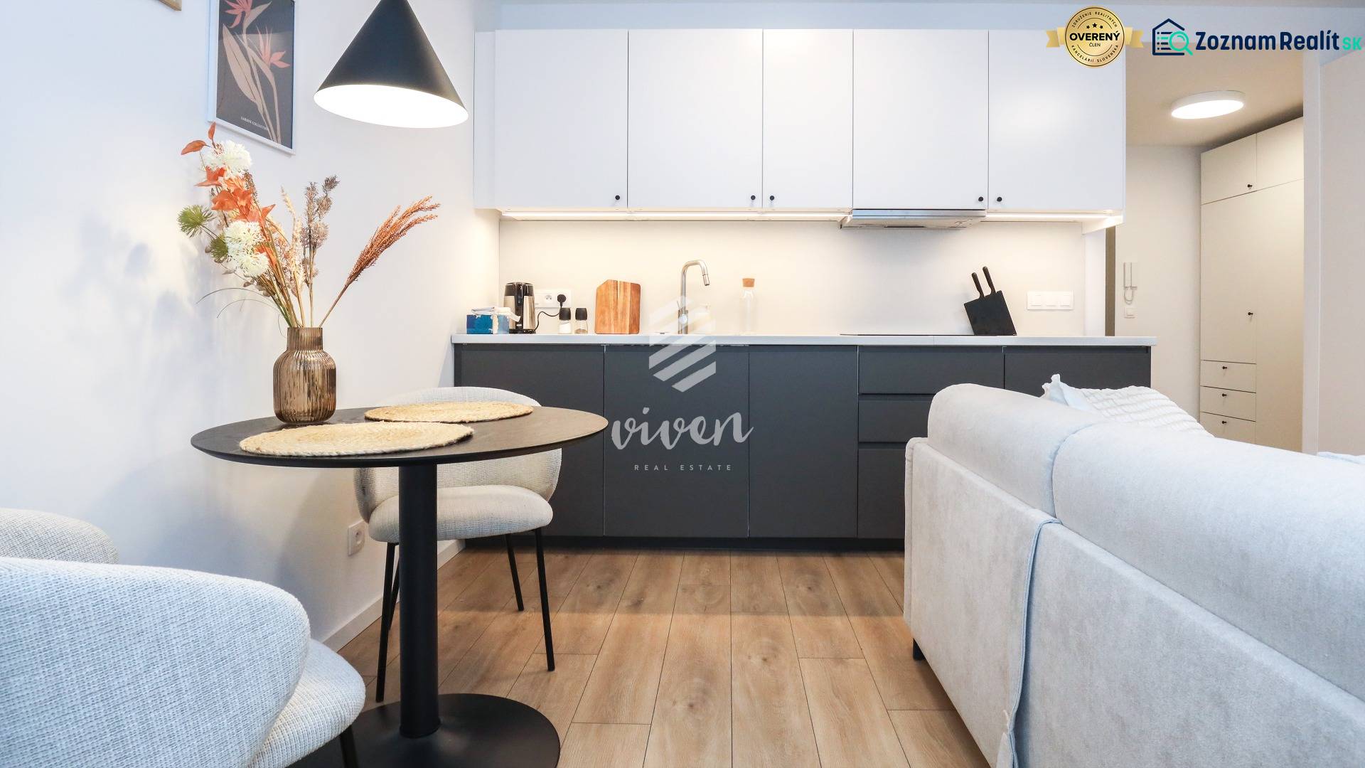 PRENÁJOM - 1 izbový byt v projekte Ovocné sady - Kuchyňa v 1-izbovom byte s bielymi skrinkami, tmavou linkou a podlahou s dreveným dekorom.