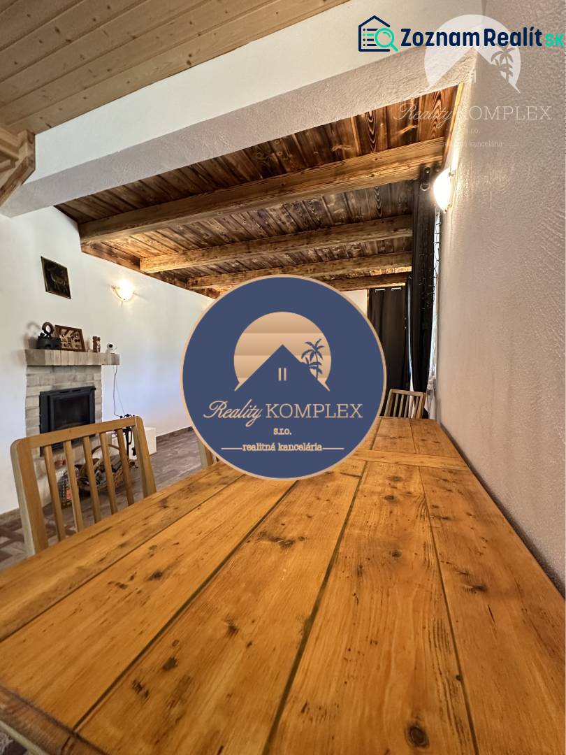 Vám ponúka na predaj rodinný dom v Komárne, ča - Interiér rodinného domu so stolom z drevom dekoru, krbom a dreveným stropom.