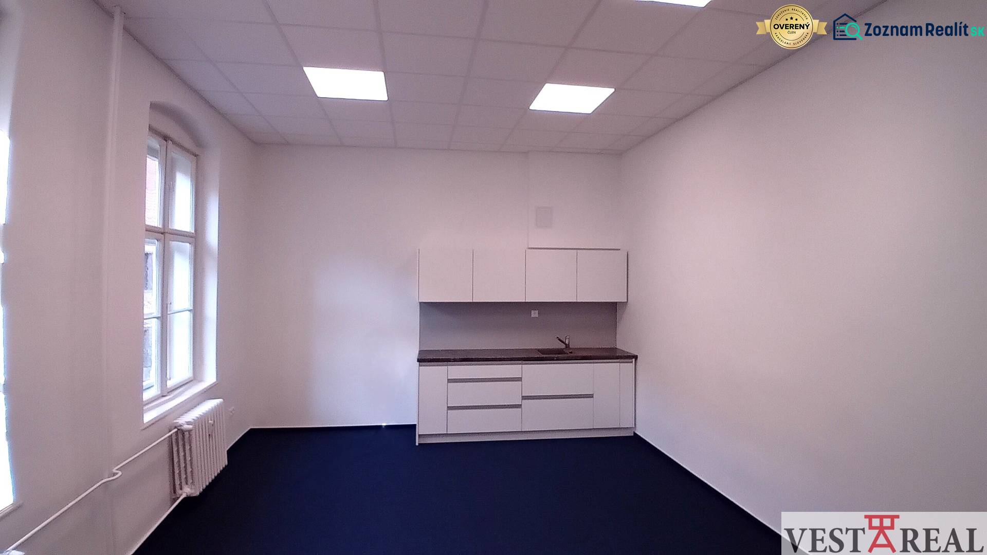 Na prenájom priestor- kancelária 60 m2 Trnava - Františkánska - Kancelárie s kuchynskou linkou a modrým kobercom, biele steny a veľké okno.