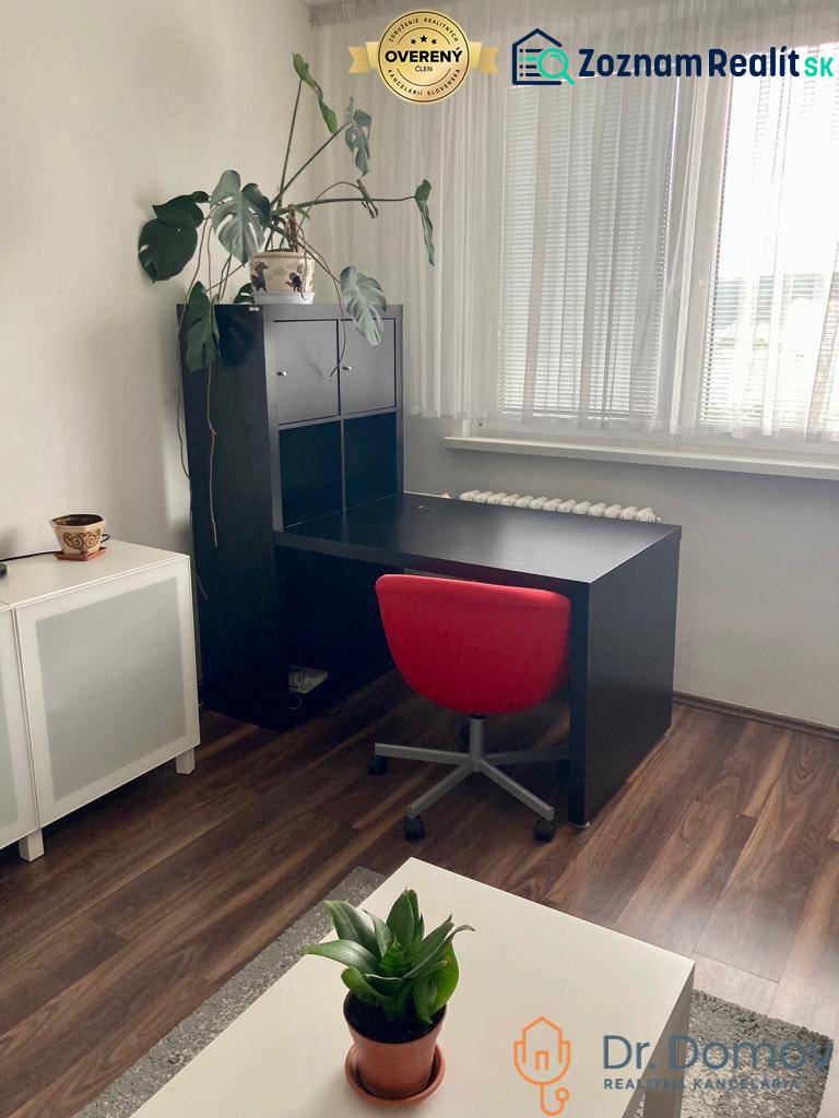NA PRENÁJOM ÚTULNÝ 1-IZBOVÝ BYT, ČIASTOČNÁ REK., KAROLA ADLERA, BA - Pracovný kút v 1-izbovom byte s čiernym stolom, červenou stoličkou a podlahou s dreveným dekorom.
