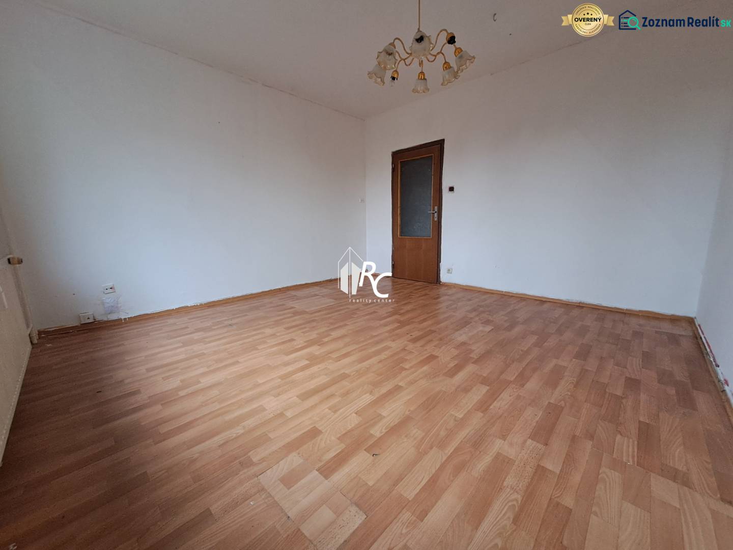 REZERVOVANÉ 2-izbový byt s nádherným výhľadom v pôvodnom stave   - Izba v 2-izbovom byte s podlahou s dreveným dekorom a lustrom.
