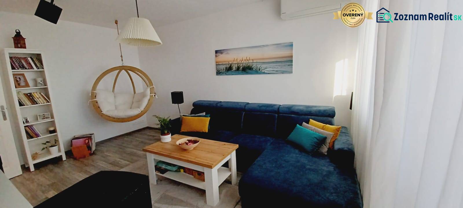 Na predaj 3izbový byt Tranovského , Dúbravka - Obývacia izba s modrou sedačkou, hojdačkou, knihovnicou a podlahou s dreveným dekorom.