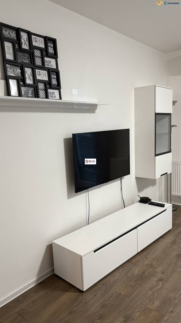 Predaj 1ibyt (garsonka) v uzavretom areáli v DNV (novostavba) - Televízor na stene, biela komoda, obrazy a podlaha s dreveným dekorom v 1-izbovom byte.