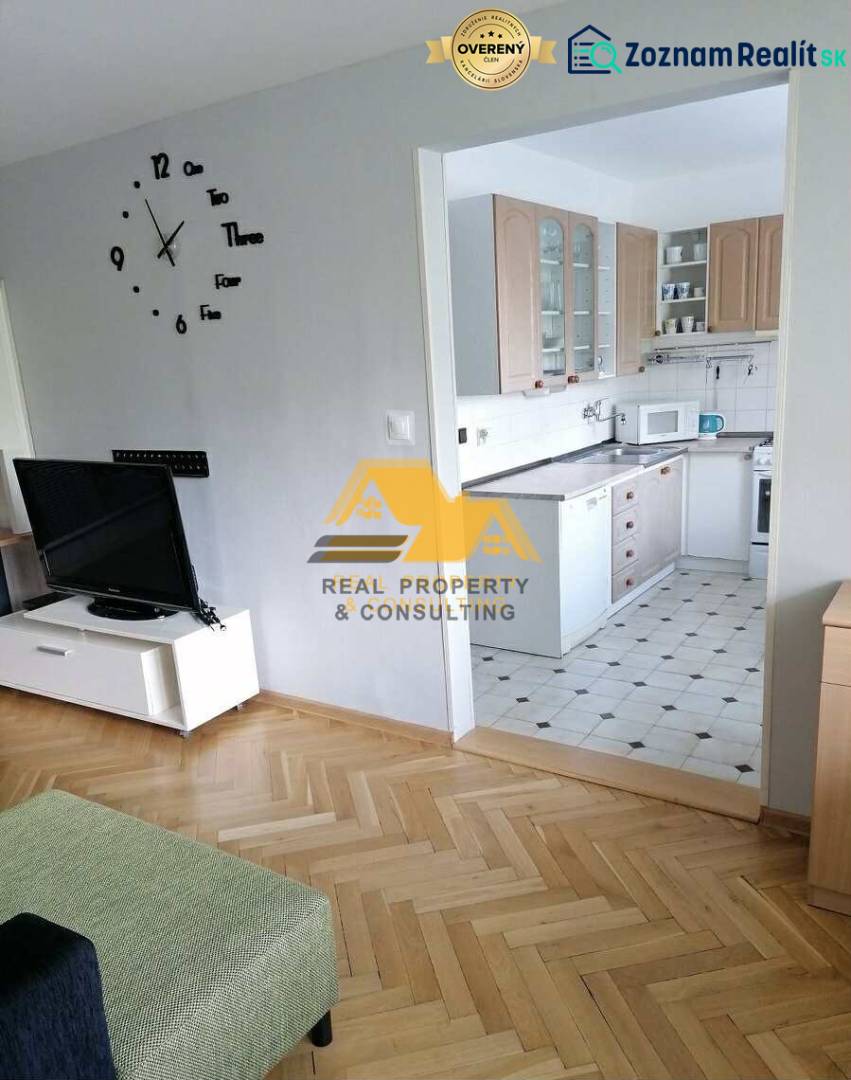 Prenajmem 3 izbový byt v Nových Zámkoch - Obývačka v 3-izbovom byte s podlahou s dreveným dekorom a kuchyňou v pozadí.