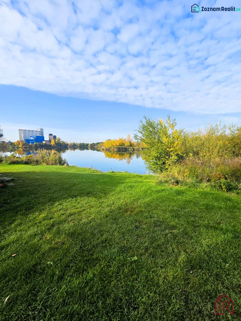 KH-1018, PREDAJ Pozemok pri jazere 887 m², garáž, altánok - Trávnatá plocha a vodná plocha obklopená vegetáciou v Čani. Výstavba domov.