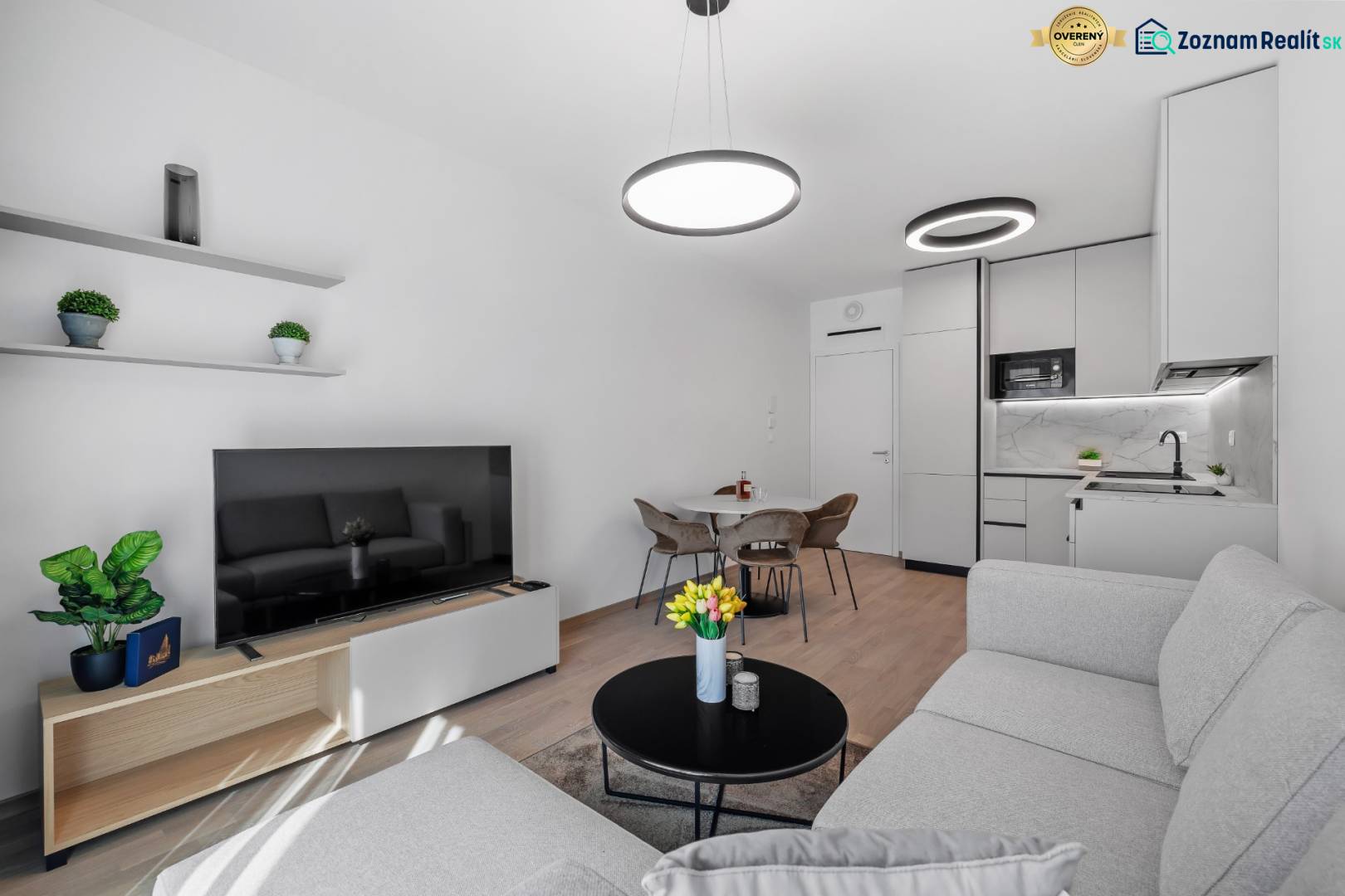 I Prenájom exkluzívneho 2-i bytu v projekte Metropolis - Obývačka s kuchyňou v 2-izbovom byte, svetlý gauč, televízor, podlaha s dreveným dekorom.