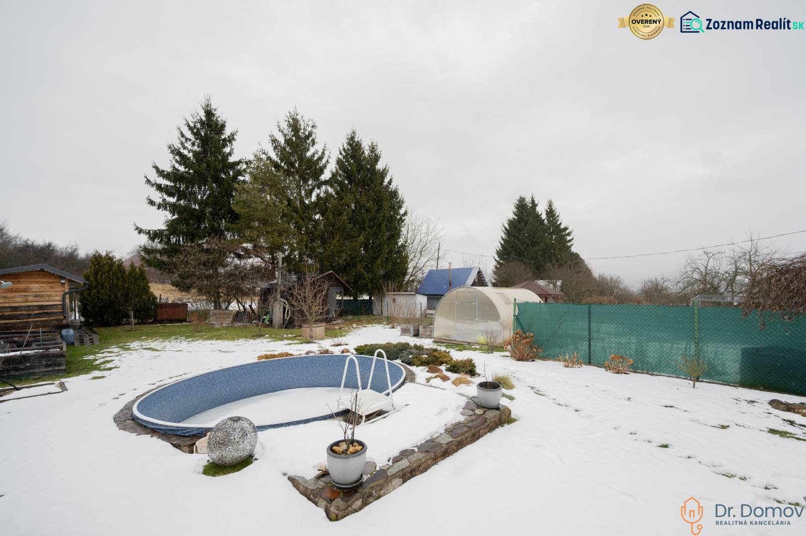 Záhradná chatka Handlová - Zasnežená záhrada pri chate v Handlovej s bazénom, skleníkmi a ihličnatými stromami.