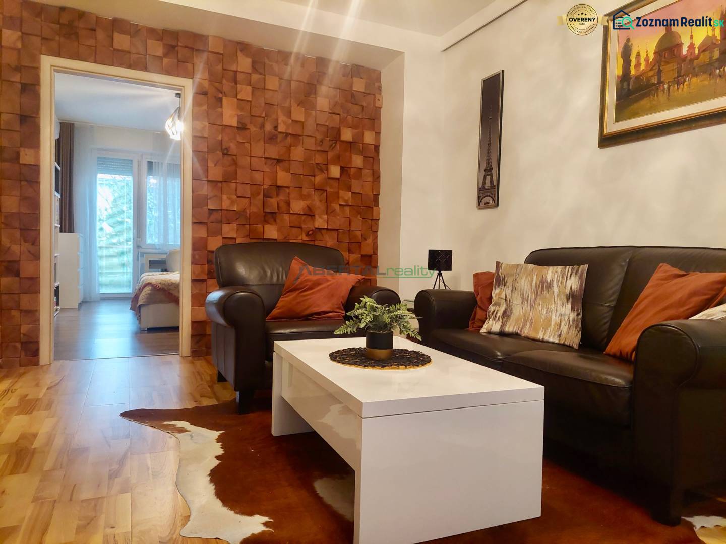 Vkusný, moderný 2 izb, klíma, 870 s ener., Sibírska ul./Nové Mesto - Obývacia izba v 2-izbovom byte s koženou sedačkou, stolíkom a stenou s dreveným dekorom.