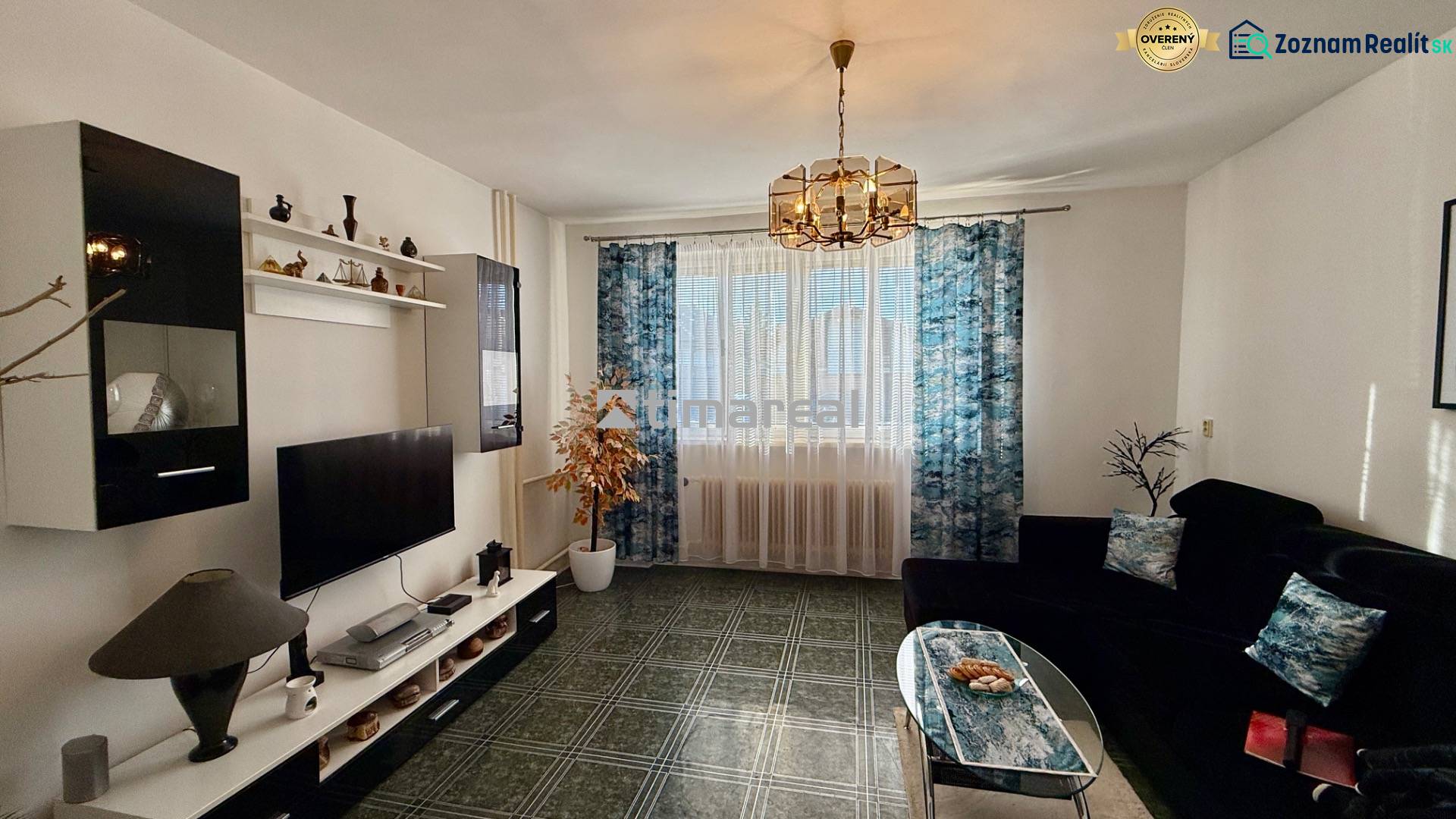 * 7i rodinný dom, veľký 33.5á pozemok, Brestovany - Obývacia izba v rodinnom dome s dekoratívnym lustrom, tmavou sedačkou, televíziou a vzorovanými závesmi.
