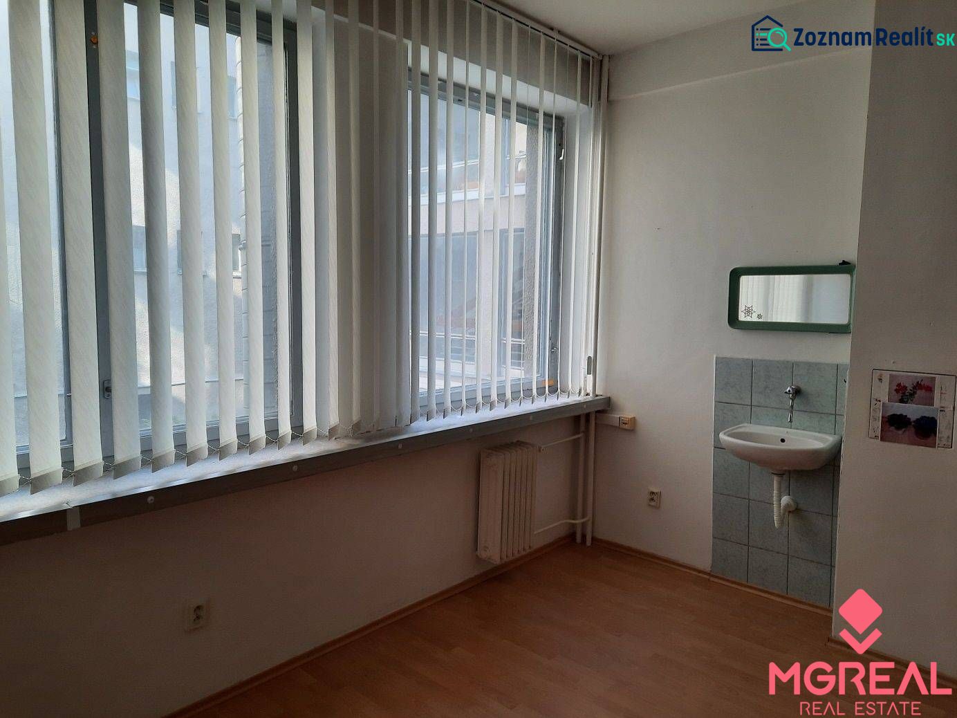 Prenájom kancelárií 21 m2 v centre Trnavy - Kancelária s umývadlom a podlahou s dreveným dekorom, svetlo cez vertikálne žalúzie.