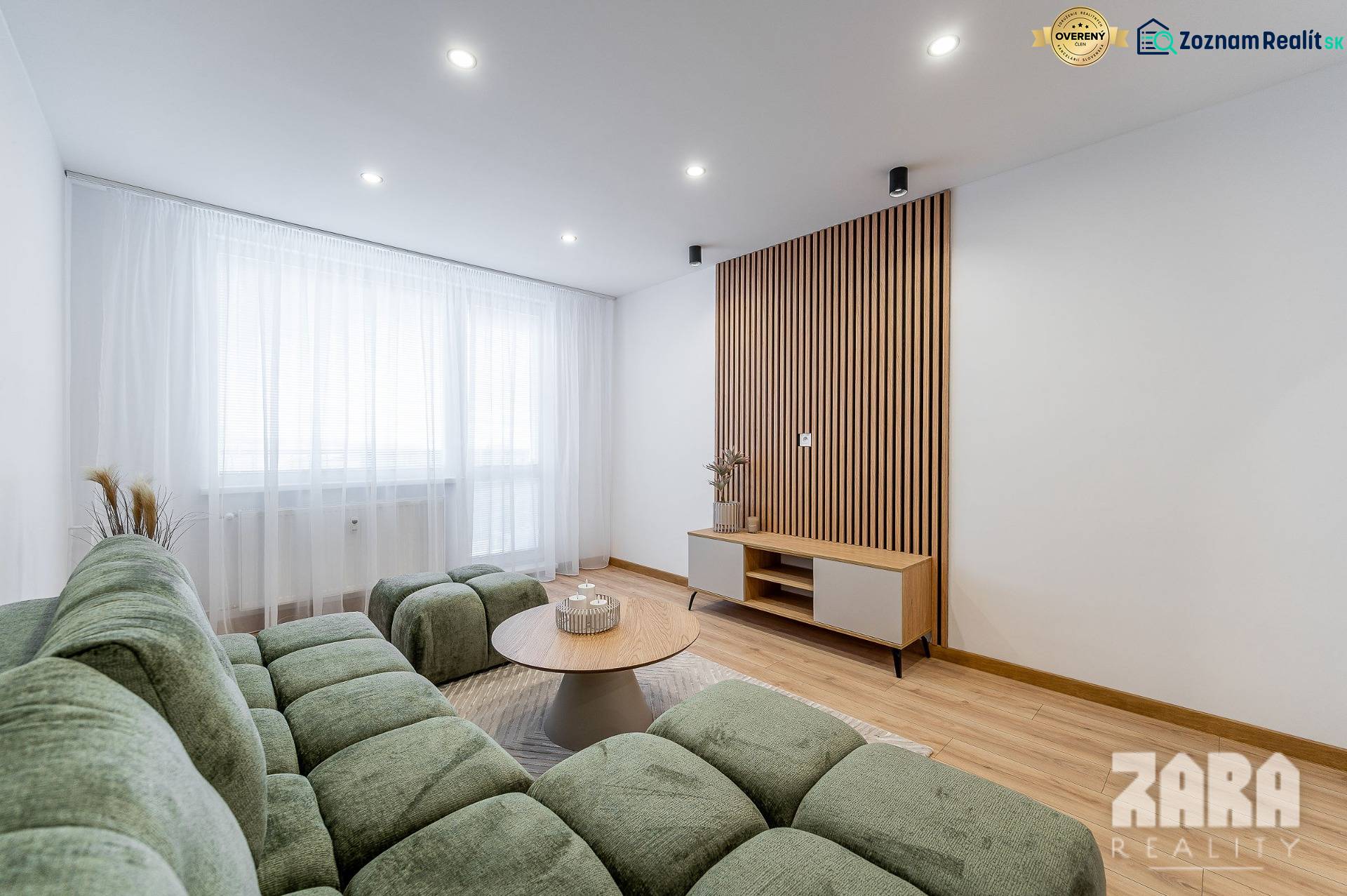 Kpt. Jaroša -  NOVINKA - MODERNÝ 2 IZBOVÝ BYT, 59 m2 + loggia. - Obývačka s podlahou s dreveným dekorom, zelenou sedačkou a svetlým nábytkom v 2-izbovom byte.