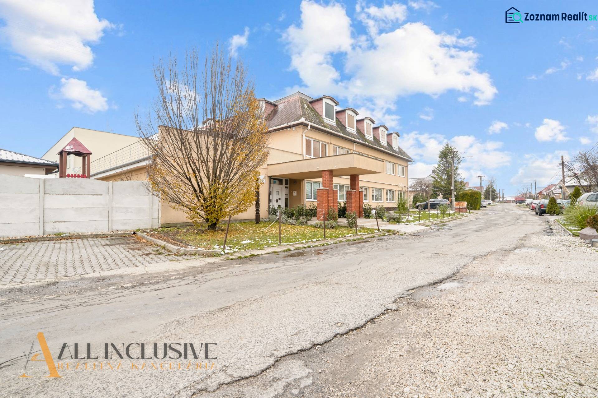 NA PREDAJ ŠKOLSKÁ BUDOVA NA VEĽKOM POZEMKU, MOSTOVÁ - Budova v Mostovej so stromom v popredí a parkoviskom na pravej strane.