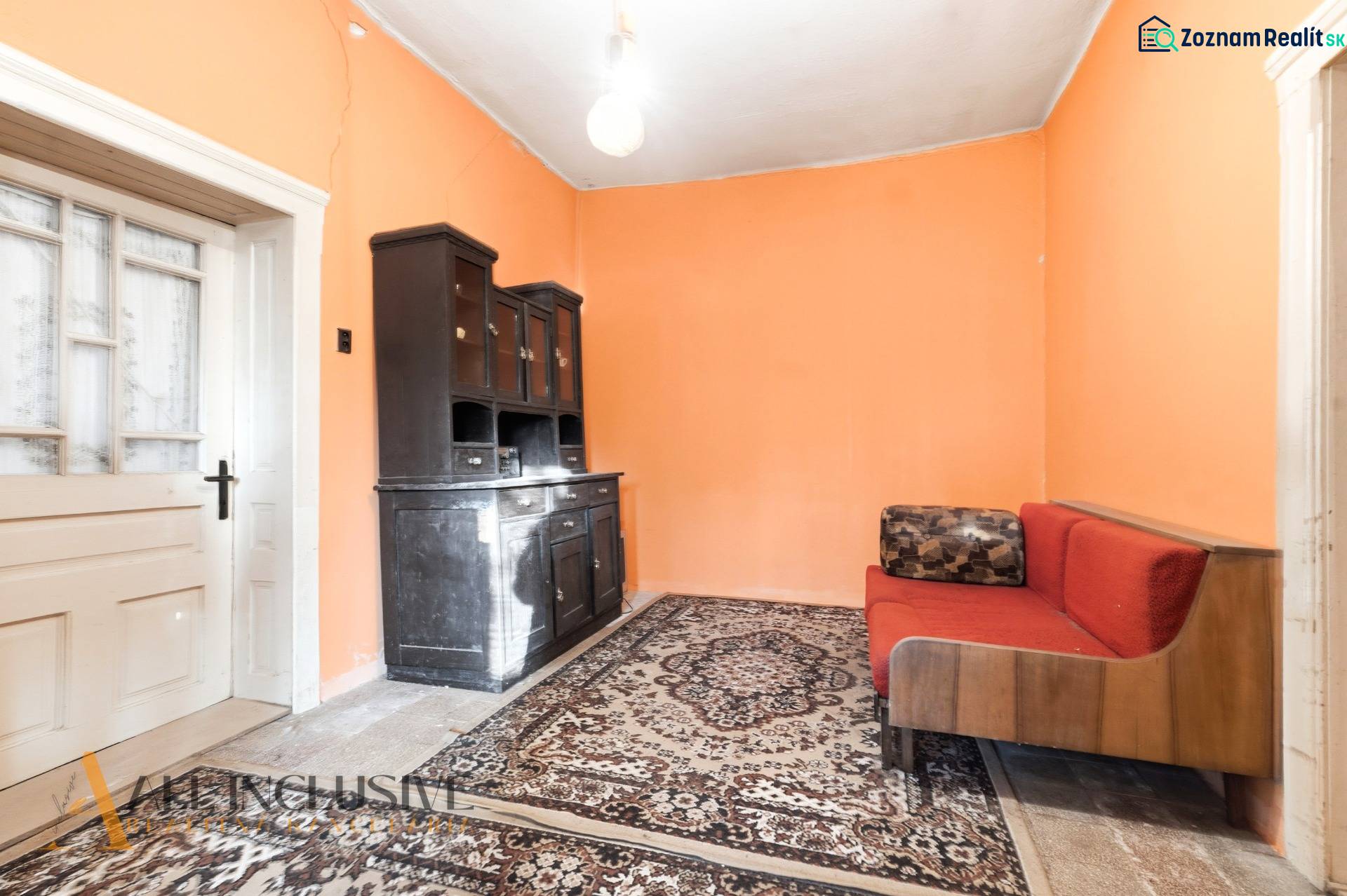 Na predaj 4-izbový rodinný dom, Dolné Saliby - Izba v rodinnom dome s oranžovými stenami, koberec, gauč a tmavá skriňa.
