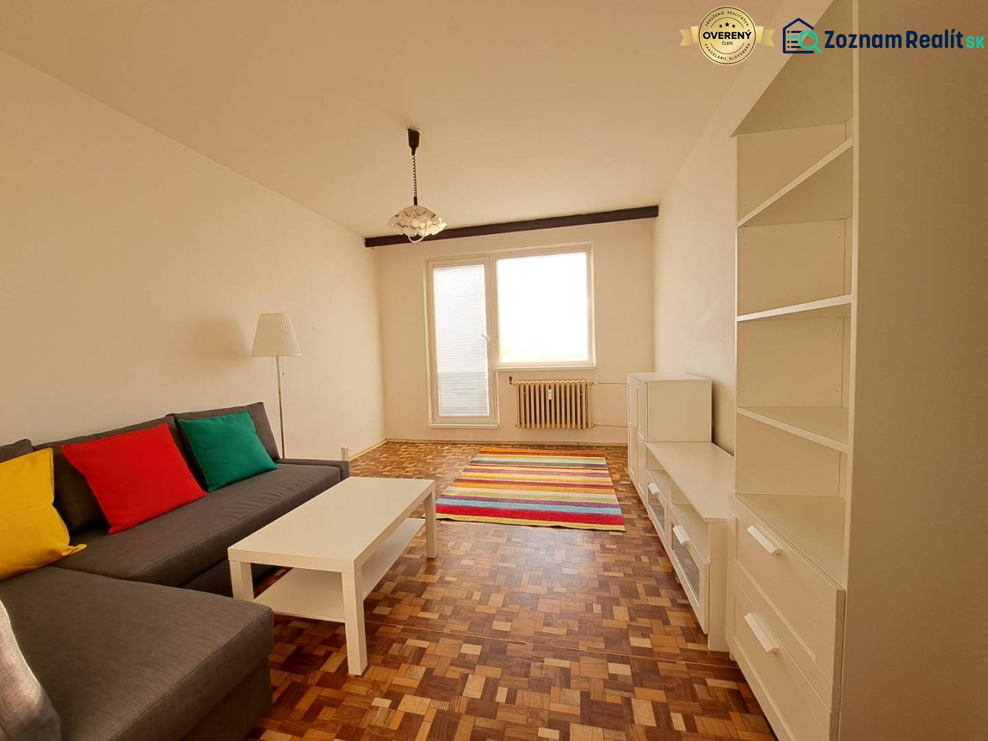 Prenájom 2-izbového bytu v Poprade na Dostojevského ul. - Obývacia izba v 2-izbovom byte s pohovkou, bielym nábytkom, pestrofarebnými vankúšmi a podlahou s dreveným dekorom.