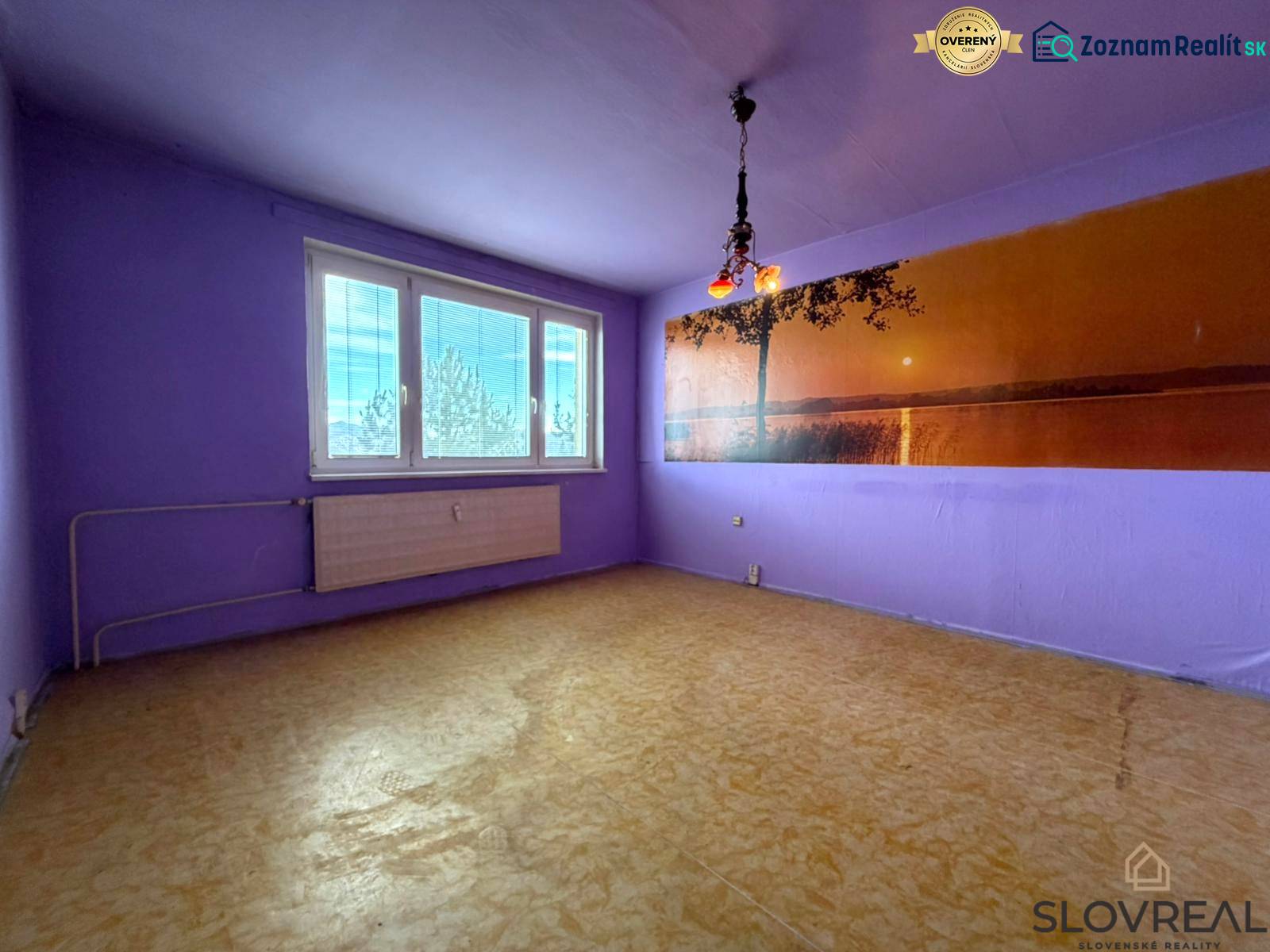 Predaj 2 - izbového bytu v Banskej Štiavnici - Fialovo ladená izba v 2-izbovom byte s podlahou s dreveným dekorom a veľkým oknom.