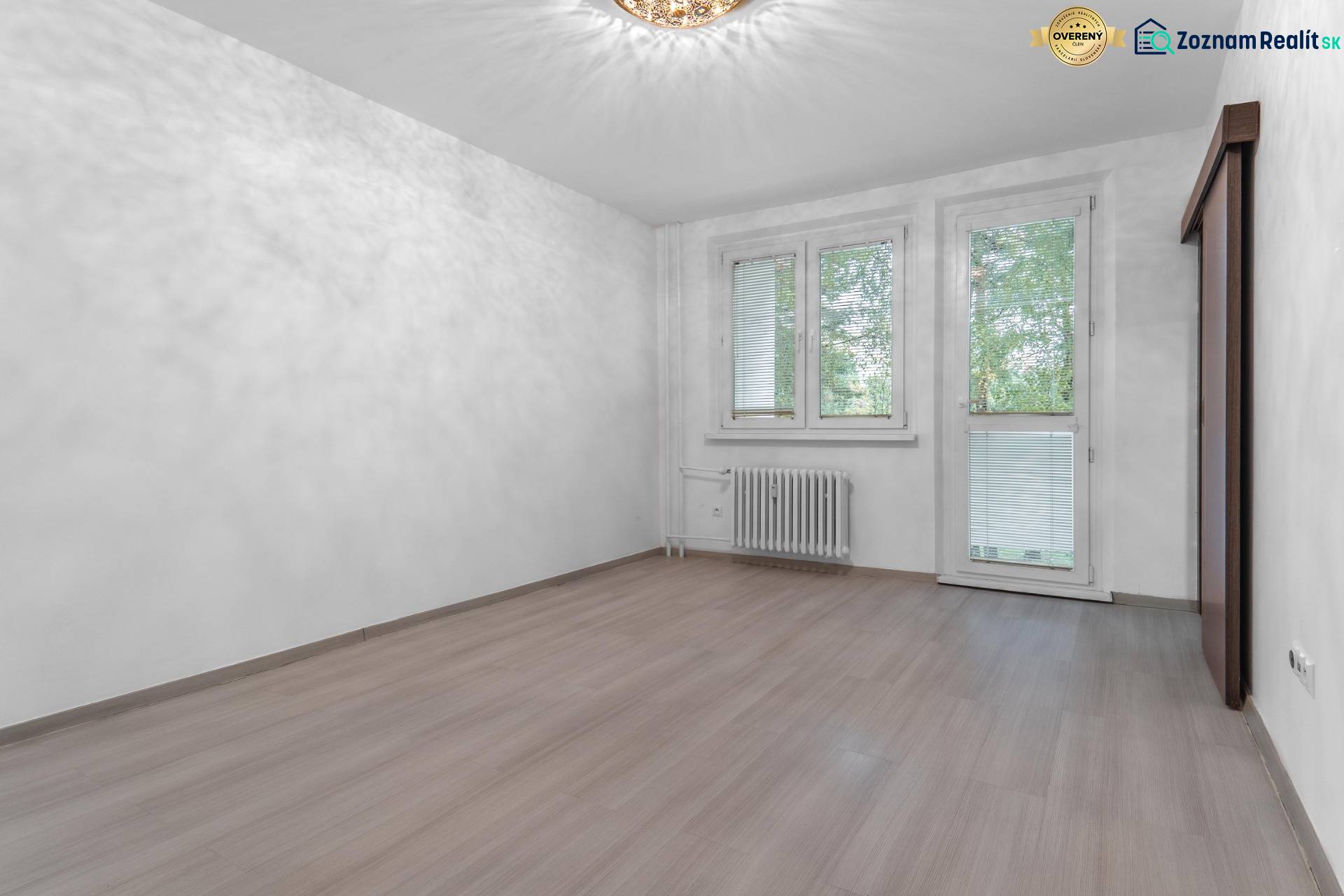 I Predaj 3- izbového bytu v Dúbravke - Izba v 3-izbovom byte s podlahou s dreveným dekorom a veľkými oknami.