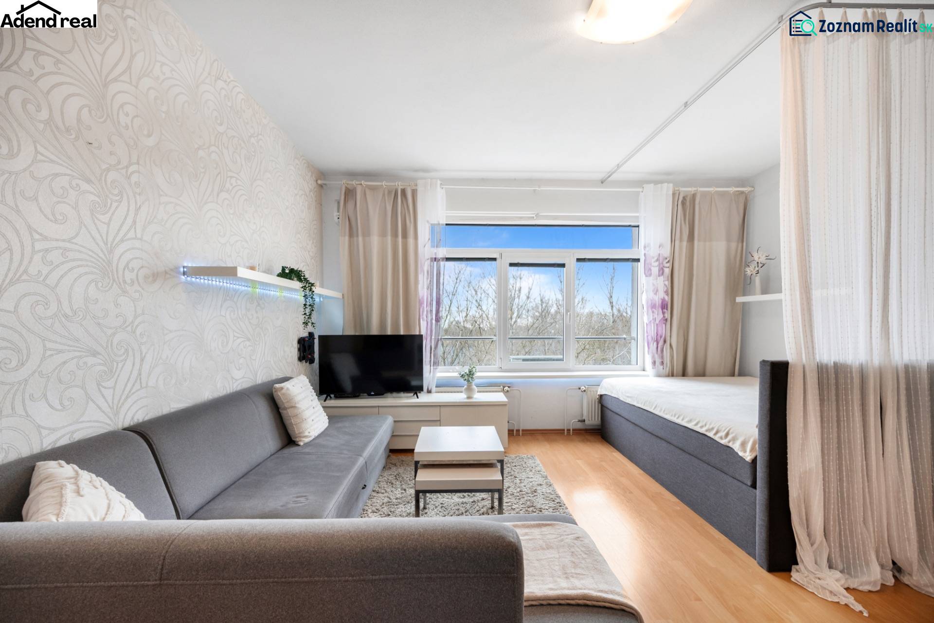 Na predaj príjemný zariadený 1 izbový byt na ulici Antolská, Petržalka - Obývacia izba s pohovkou, závesom a televíziou, podlaha s dreveným dekorom.