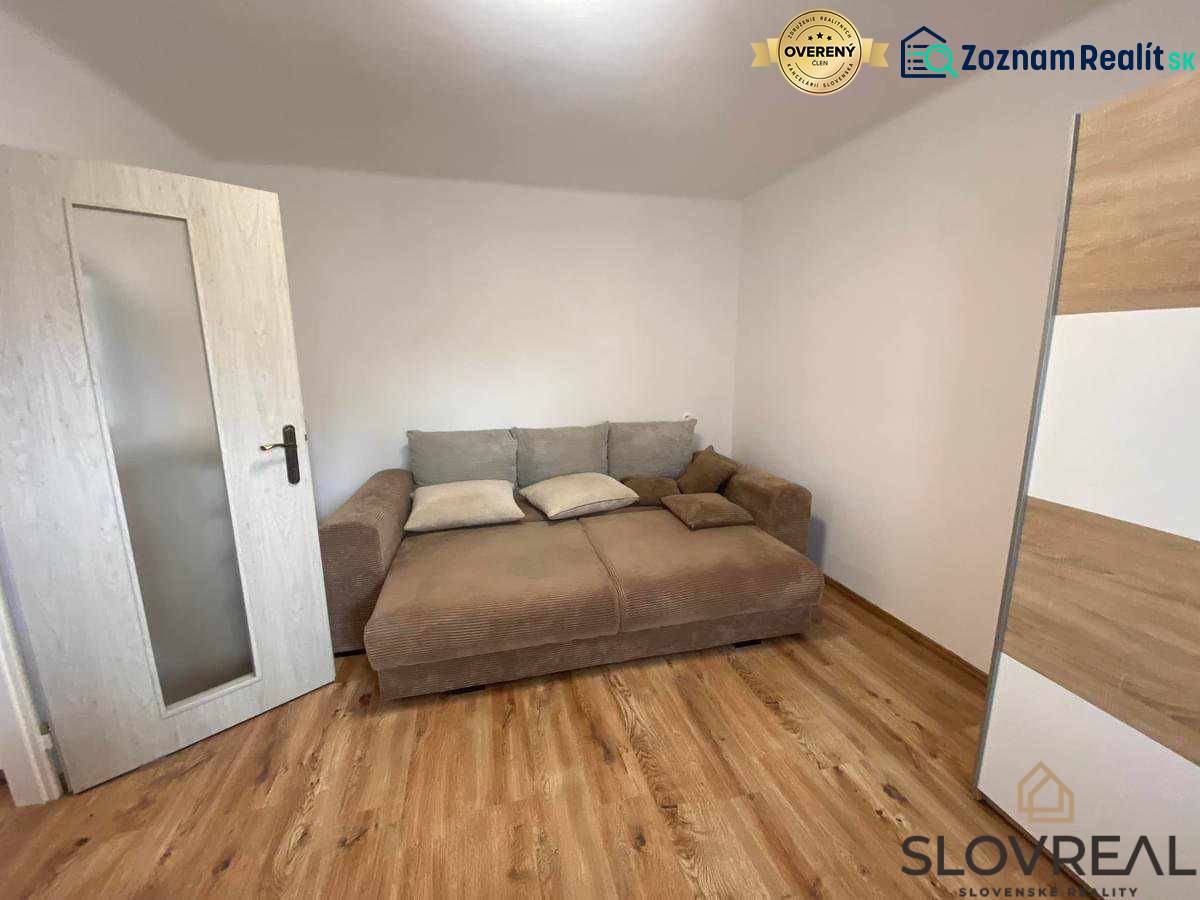 Predaj 3 - izbového bytu - Nová Baňa - Izba s hnedou pohovkou, bielymi stenami a podlahou s dreveným dekorom v 3-izbovom byte.