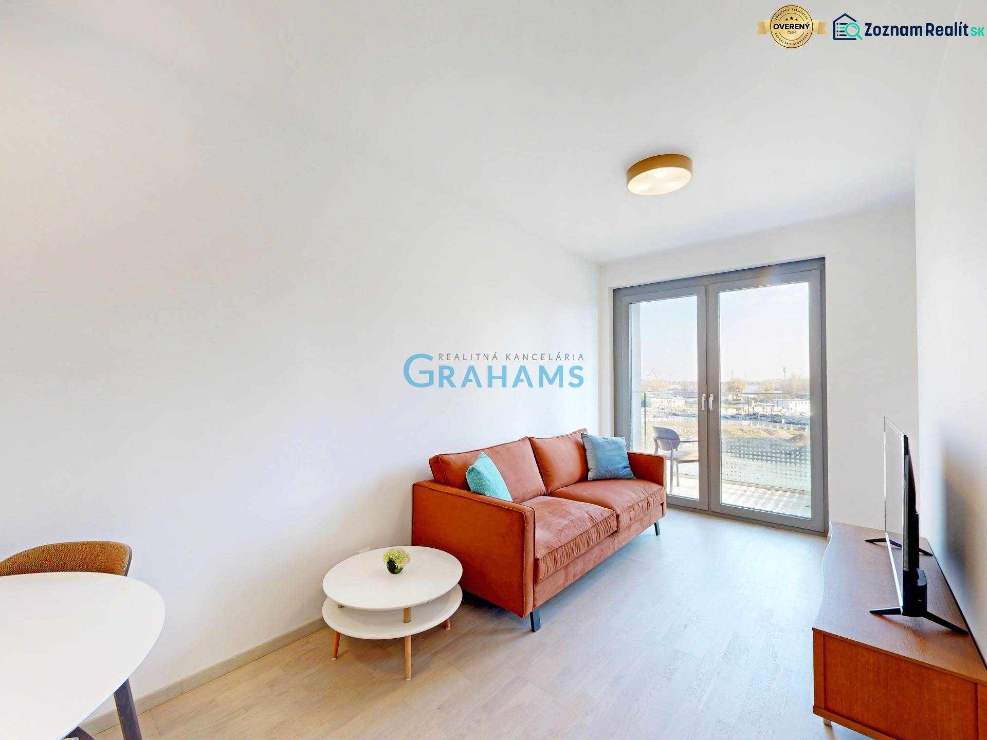 Nový, ešte neobývaný 1,5-izbový byt, parkovanie, Metropolis, Bottova - Obývacia izba v 2-izbovom byte s oranžovou pohovkou, TV a balkónovými dverami.