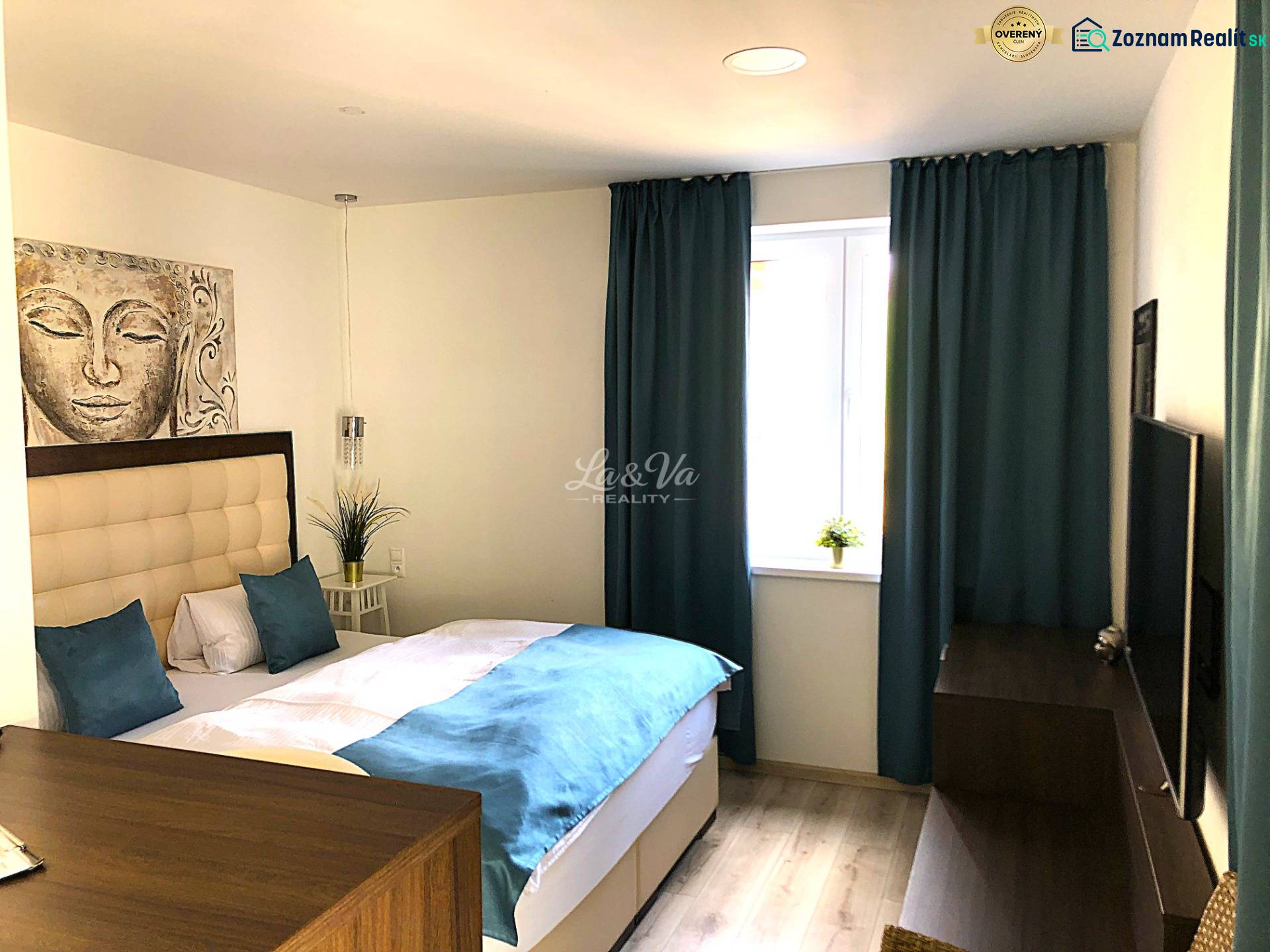 Na predaj apartmán s veľkou terasou Podhájská. - Garsónka so svetlým nábytkom, posteľou a modrými závesmi, podlaha s dreveným dekorom.
