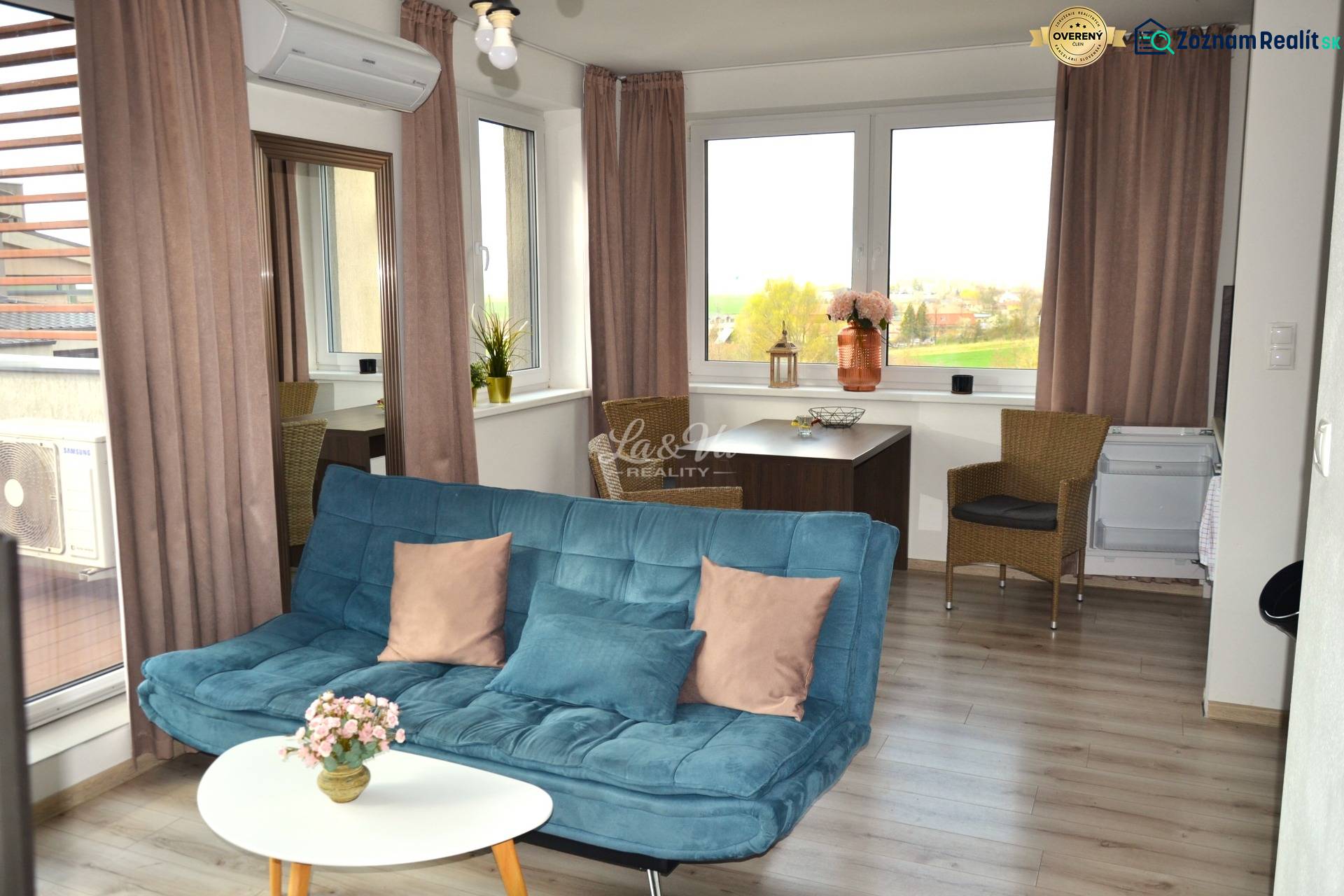 Na predaj veľký 49m2 apartmán Podhájská.  - Obývacia izba so sedačkou, stolíkom, rastlinami a podlahou s dreveným dekorom v 2-izbovom byte.