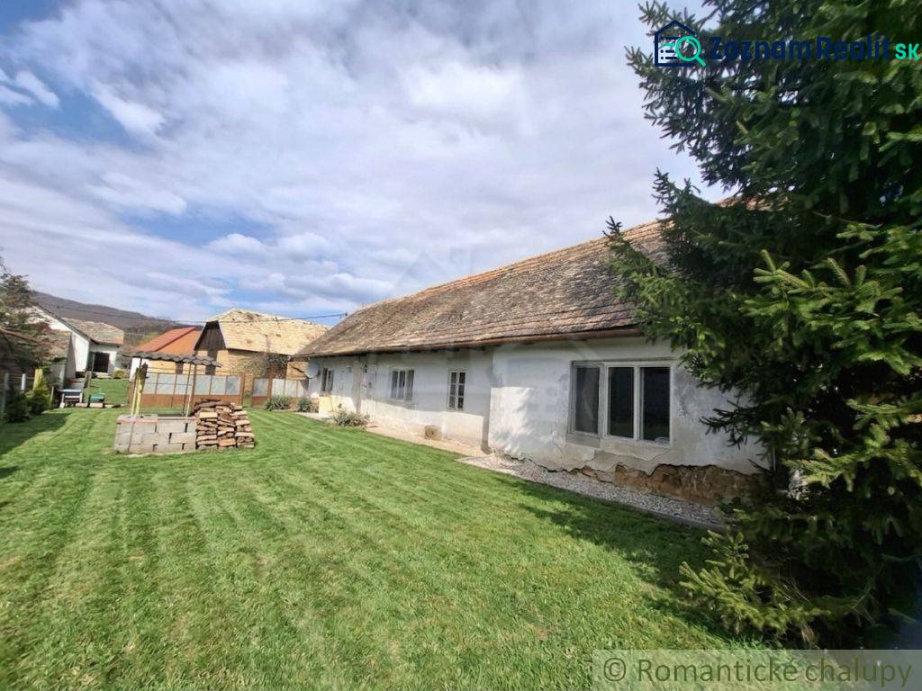 Vymeňte mestský hluk za ticho Bystričky: Domček s dušou a potenciálom - Rodinný dom v Ozdíne s pôvodným vzhľadom a trávnatým dvorom, obklopený stromami.