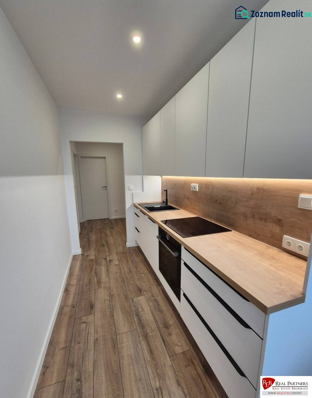 Prenájom 2-izbového bytu Bratislava II, Ružinov - Úzka kuchyňa v 2-izbovom byte s drevenou podlahou a bielymi skrinkami.