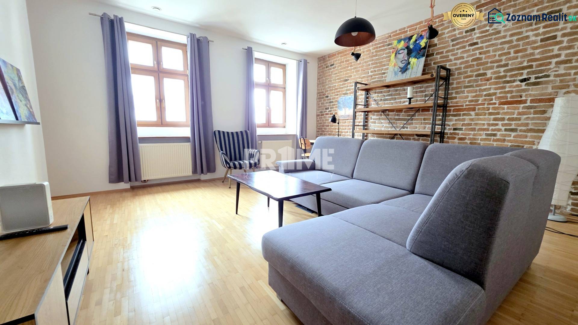 JEDINEČNÝ 2i byt, REKONŠT., 2 X .SAMOST.IZBA, Hviezdoslavovo nám.  - Obývacia izba s tehlovou stenou, sivou sedačkou a podlahou s dreveným dekorom v 2-izbovom byte.