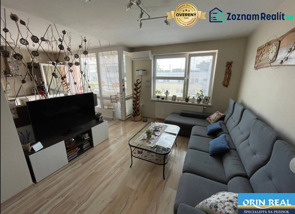 PREDAJ PRIESTRANNÉHO 3 IZB. BYTU NA MUŠKÁTE S GARÁŽOVÝM STÁTIM - Obývacia izba s podlahou s dreveným dekorom, šedou sedačkou a televízorom v 3-izbovom byte.