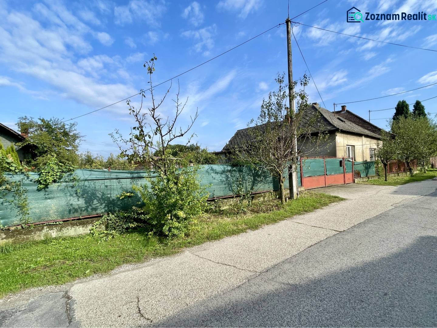 JAHODNÁ – STARÝ RODINNÝ DOM S PEKNÝM STAVEBNÝM POZEMKOM NA PREDAJ - Rodinný dom v Jahodnej s oplotením a stromami popri neuvedenej ulici za slnečného dňa.