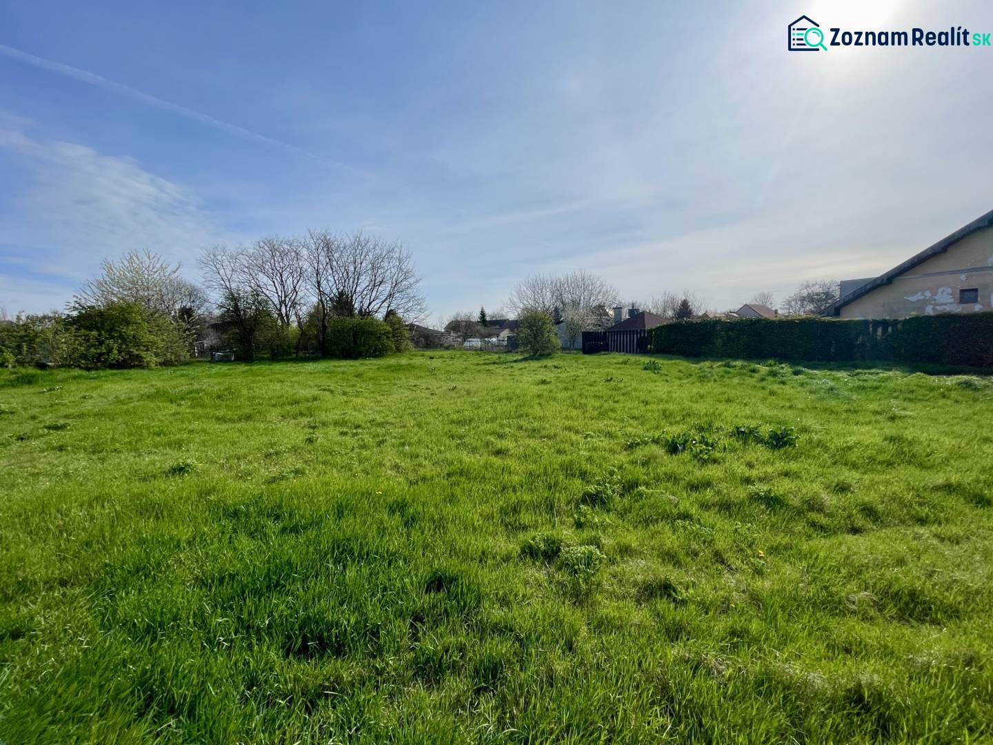 OKOČ – POZEMOK NA PREDAJ V CENTRE DEDINY - Pozemok na bývanie v Okoči s trávnatou plochou a stromami v pozadí.