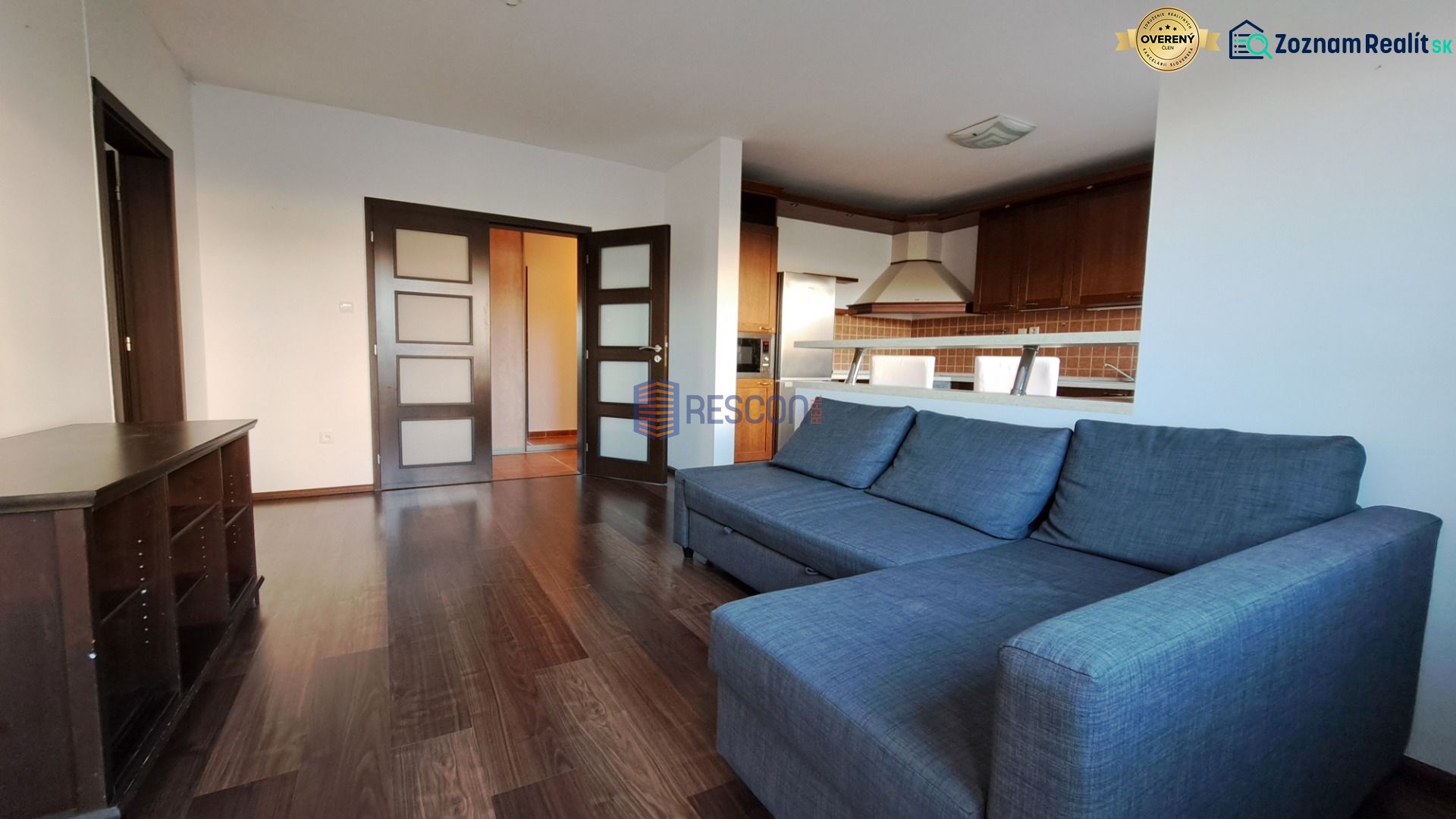 Bývanie s vlastnou predzáhradkou – zariadený 2-izbový byt (62+33 m²) - Obývacia izba v 2-izbovom byte s podlahou s dreveným dekorom a kuchynským kútom.