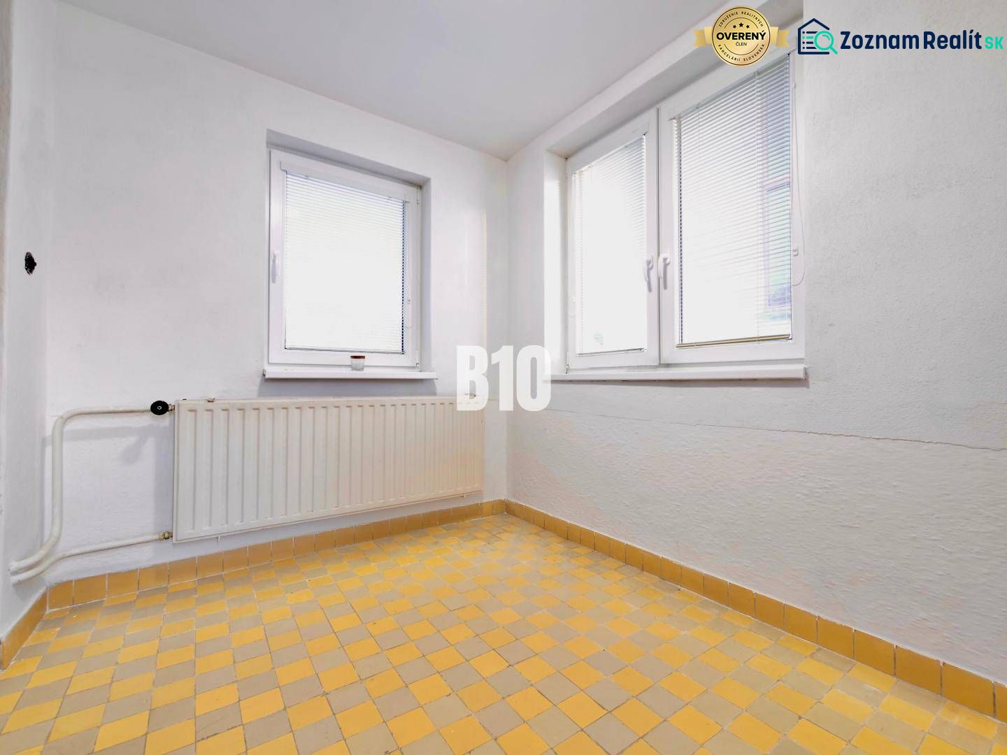 Rodinný dom s rovinatým 10a pozemkom - Biela stena, okná, radiator a žltá kachľová podlaha v rodinnom dome.