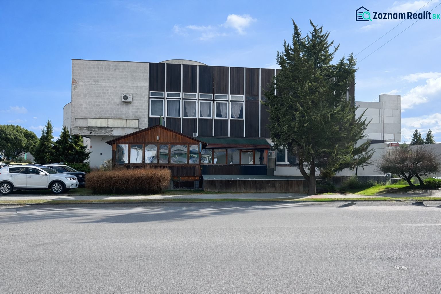 Komerčné priestory pri hlavnej ceste s parkoviskom -Žiar nad Hronom - Exteriér gastro priestorov v Žiari nad Hronom, obklopený stromami a zaparkovanými autami.