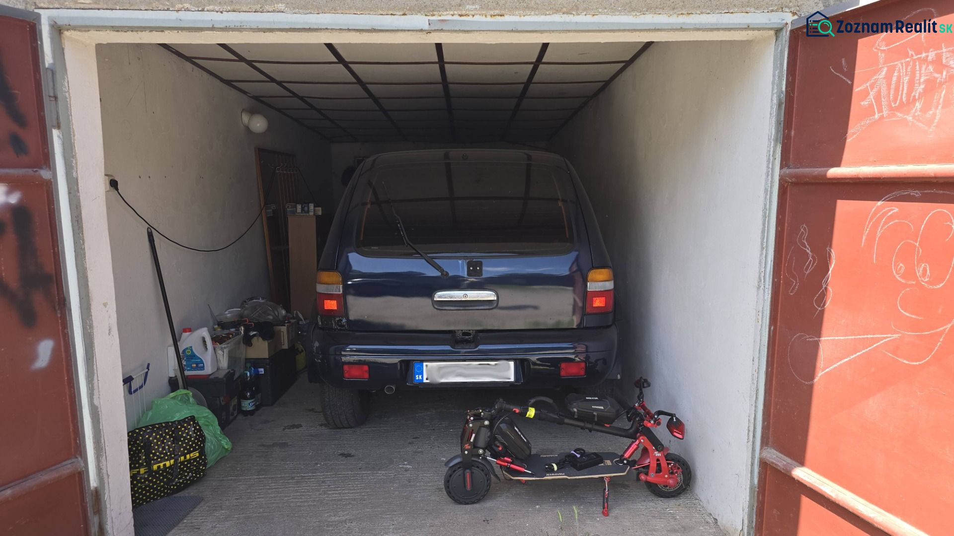 NA PREDAJ MUROVANÁ GARÁŽ 18 m² VO ZVOLENE – ELEKTRINA, VLASTNÝ POZEMOK - Garáž s autom a elektrickou kolobežkou, uloženými predmetmi a kresbou na dverách.