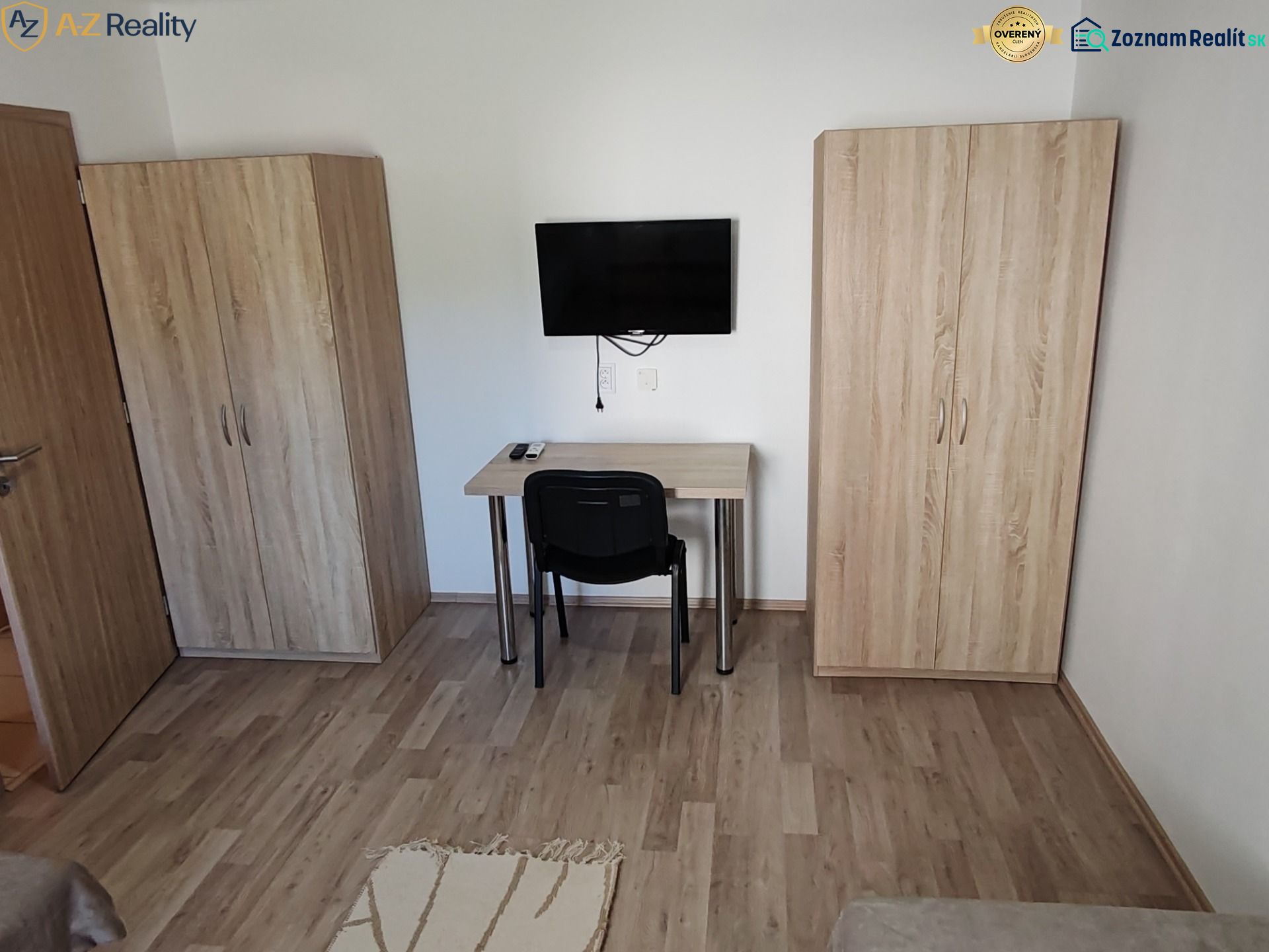 Na prenájom 1 izbový byt v Komárne - Izba s podlahou s dreveným dekorom, televízorom, stolom a dvomi skriňami v 1-izbovom byte.