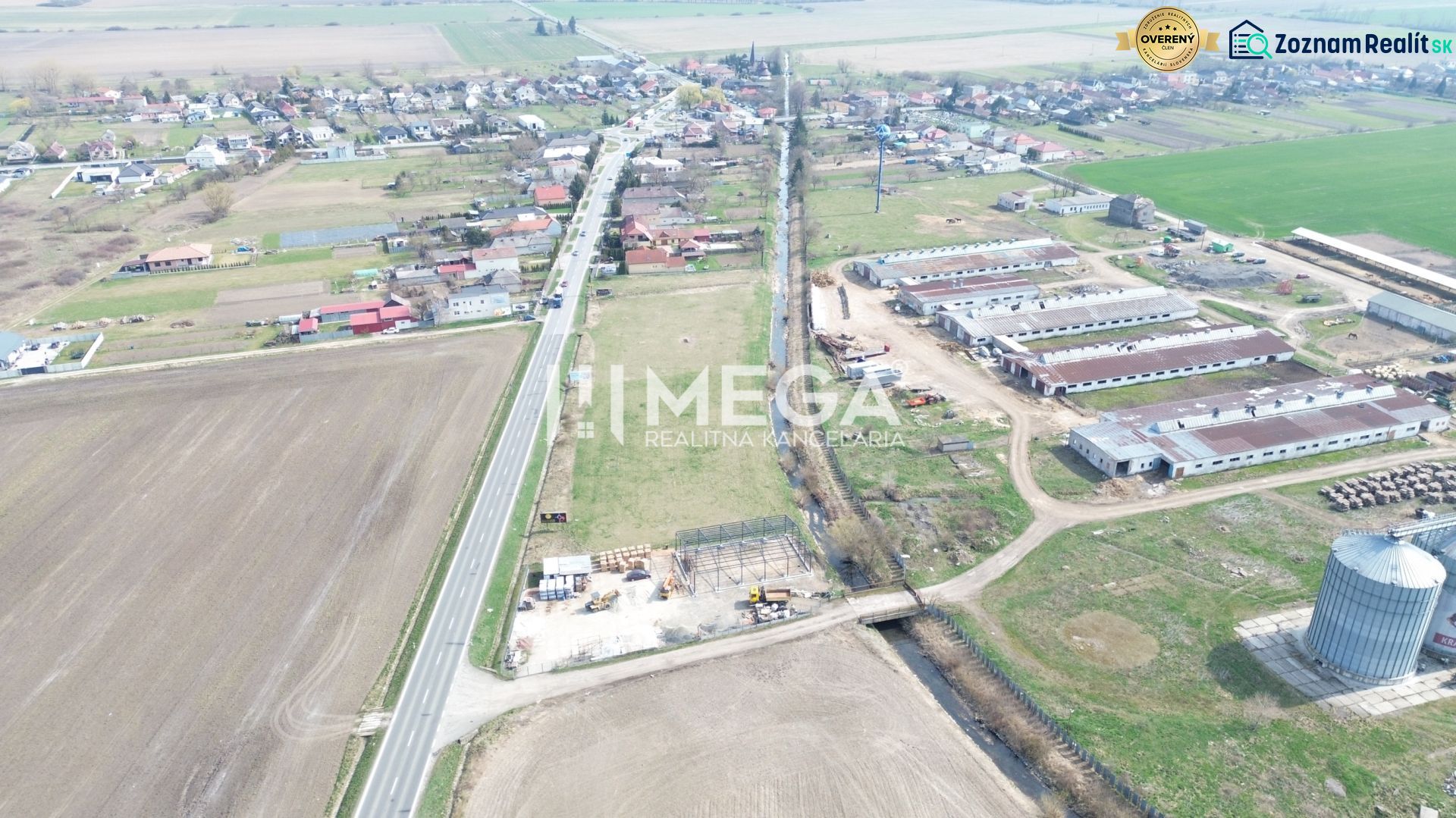 NA PREDAJ EXKLUZÍVNE: Lukratívny pozemok 6 086m2 – Hriadky (okres TV)  - Letecký pohľad na výrobné priestory v obci Hriadky obklopené poľnohospodárskymi políčkami a cestou.