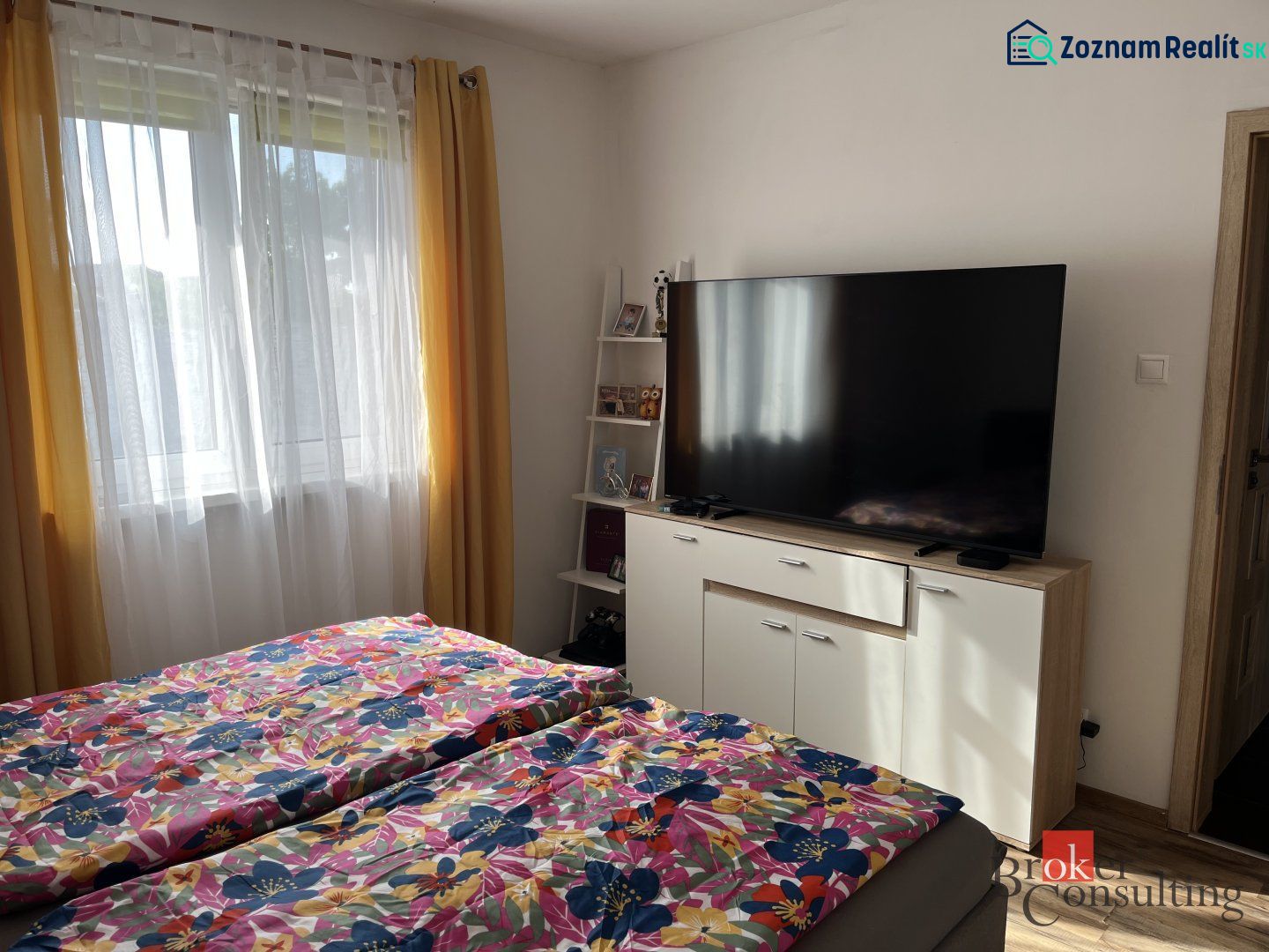 Predaj bungalovu - drevodom - Spálňa v rodinnom dome s TV, kvetinové obliečky, žlté závesy, okno.