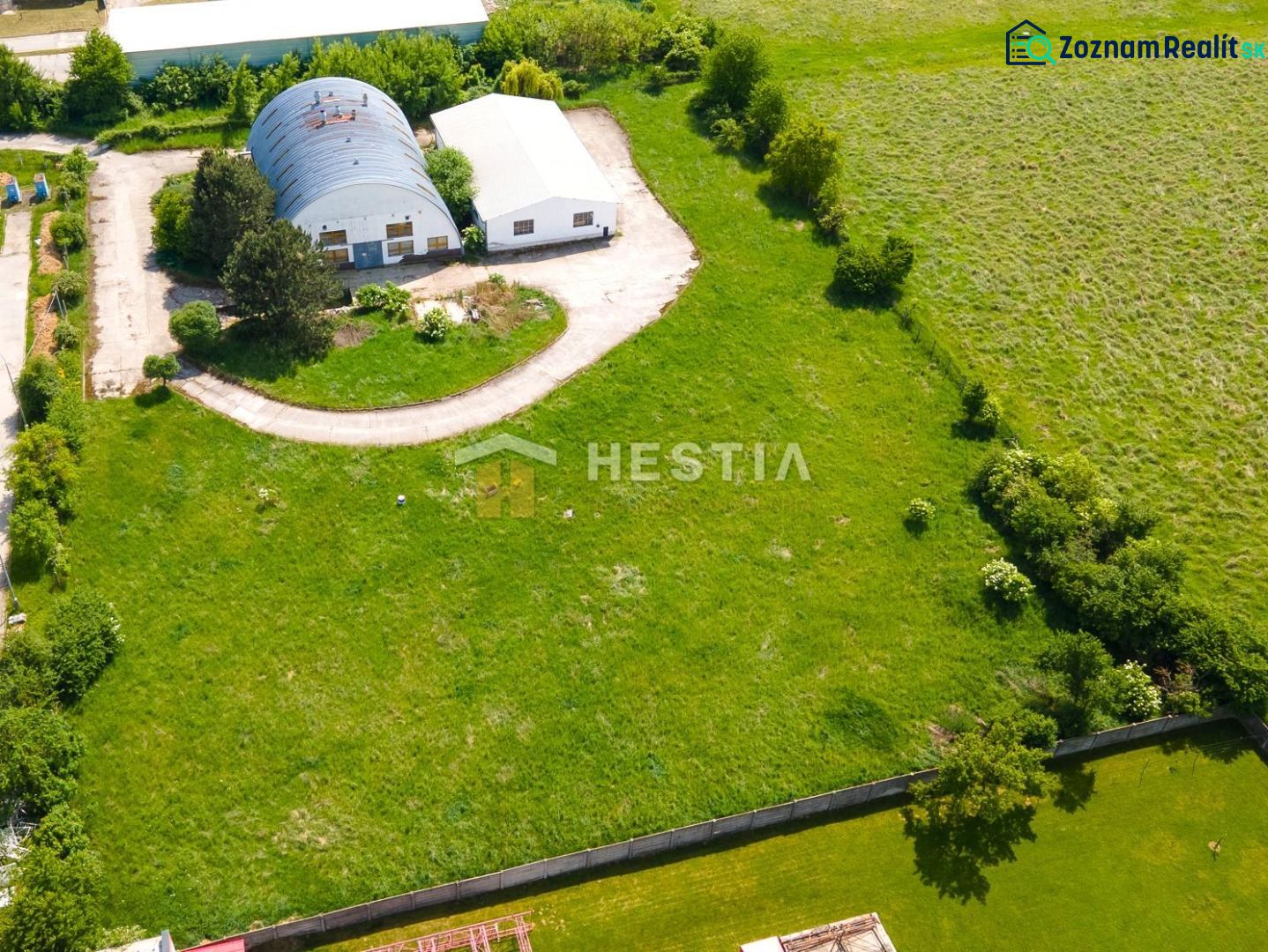 Skladový areál s pozemkom 10 000 m2 v priemyselnej zóne - Letecký pohľad na sklady a haly v Senici obklopené zeleňou a prístupovou cestou.