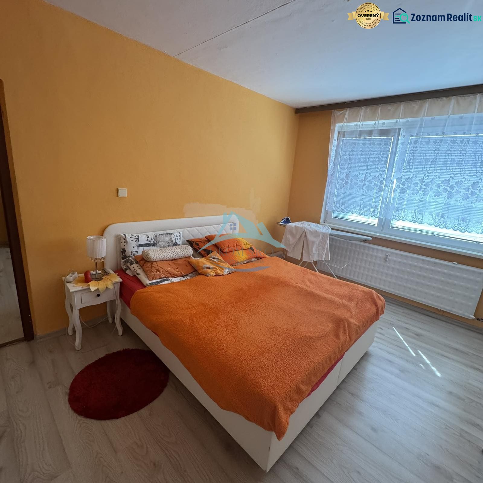 Váš nový štart alebo bezpečná investícia v srdci Šiah - Spálňa s oranžovou stenou, posteľou a podlahou s dreveným dekorom v 2-izbovom byte.
