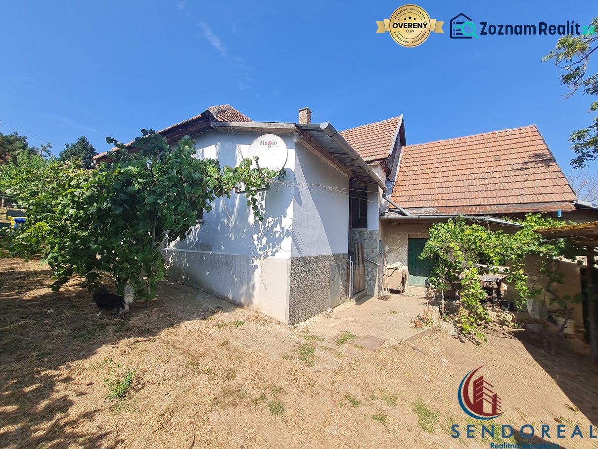 RD Otrokovce 1071m pozemok - Rodinný dom v Dolných Otrokovciach s bielou omietkou a škridlou, zelené rastliny na záhrade.