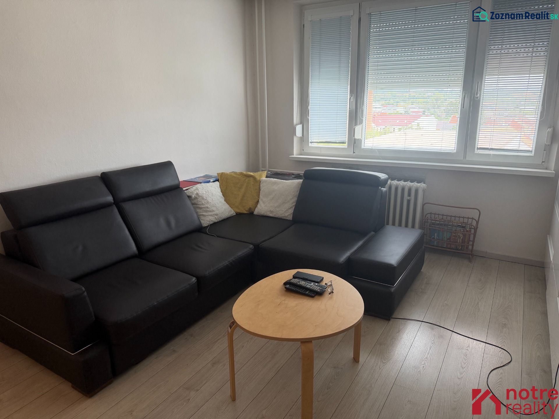 2,5 izbový byt v centre Lučenca - Obývacia izba v 2-izbovom byte s čiernym gaučom a podlahou s dreveným dekorom.