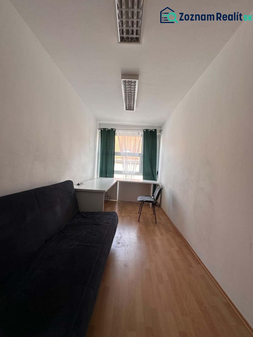 Na prenájom menšia  kancelária v centre mesta DS - 12,94 m2 - Úzka kancelária s pohovkou, stolom a stoličkou; podlaha s dreveným dekorom.