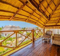ZARIA GARDEN BUNGALOWS - Drevená terasa s prútenými kreslami, výhľad na tropickú prírodu, výstavba domov v pozadí.