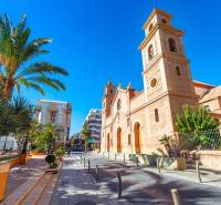 TORREVIEJA - UVÁDZACIE CENY ZA NOVOSTAVBU len 300 metrov od mora - Ulica Campoamor 12 v Torrevieji s kostolom a palmami za slnečného dňa.