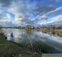 Priestranný pozemok so starším rodinným domom v obci Tvrdošovce - Idylická krajina s jazerom, oblakmi a rodinnými domami v pozadí pri brehu.