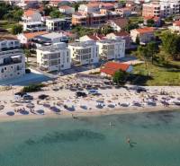 CHORVÁTSKO - Luxusné apartmány (4i) - ostrov VIR - Pláž s lehátkami a slnečníkmi, za ňou domy a budovy v letovisku.